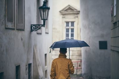 Rear view of man with umbrella walking against buildings during rainy season