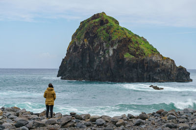 Rear view of woman standing on rocks by sea against sky