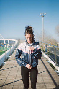Young woman in jacket standing on footbridge against sky