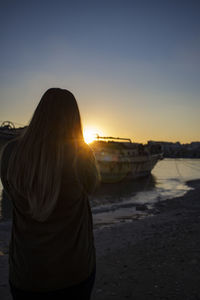 Rear view of woman standing on shore against sky during sunset