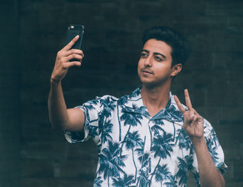 Smiling young man gesturing while taking selfie through mobile phone