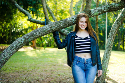 Portrait of happy young woman standing by tree trunk at forest