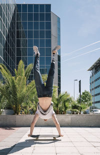 Sportive young man with artificial leg performing handstand on skateboard on pavement on sunny day on city street