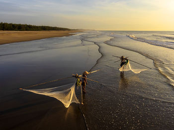 Fishermen with fishing nets standing at beach against sky during sunset