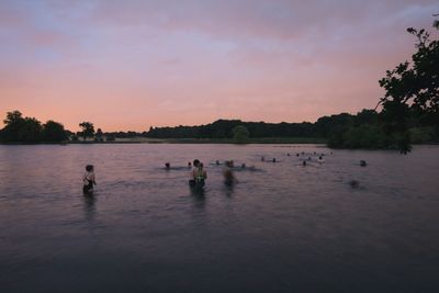 People swimming in lake against sky during sunset
