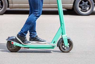 A man in blue jeans rides a rented electric scooter on a sunny day with a passing car .