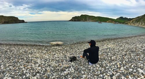 Man sitting on pebbles at beach against sky