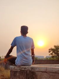 Rear view of man sitting on retaining wall against orange sky