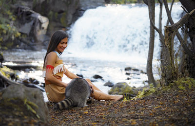 Smiling young woman in traditional clothing sitting by raccoon in forest