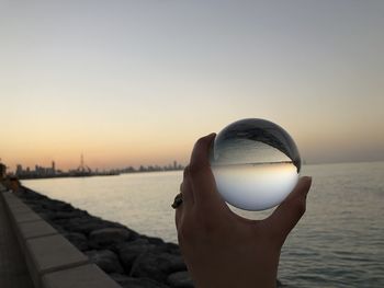 Person holding crystal ball against sky during sunset