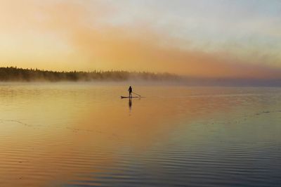 Silhouette person paddleboarding in river against sky during foggy weather at sunset