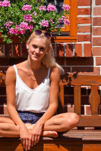 Portrait of young woman sitting on bench against brick wall during sunny day