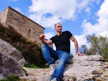 Low angle portrait of man sitting on rock against sky