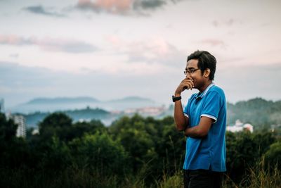 Young man looking away while standing in forest against sky