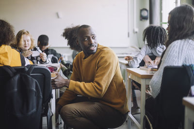 Side view of male teacher talking with student while crouching amidst desks in classroom