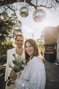 Happy bride holding bouquet standing with groom under disco balls on sunny day