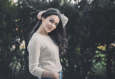 Portrait of smiling young woman with hand in hair standing against trees