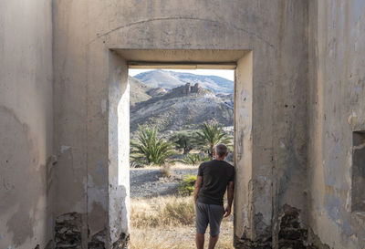 Rear view of adult man standing in doorway of an abandoned building looking at view