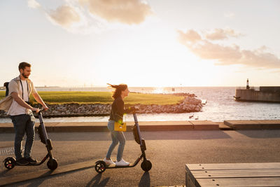 Full length of couple enjoying while riding electric push scooters on road by sea against sky