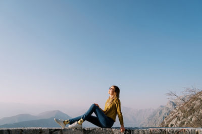 Rear view of woman sitting on rock against clear sky
