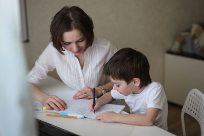 Beautiful mother teaches her son at home at the table, real interior