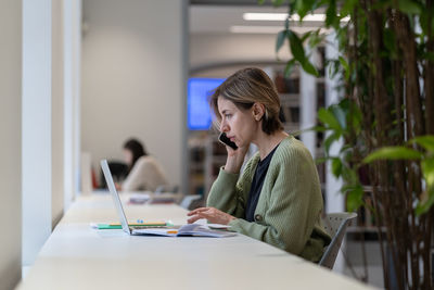 Young focused female freelancer talking on mobile phone while working remotely on laptop in library