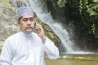 Thoughtful man talking on phone while sitting against waterfall