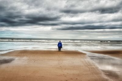 Rear view of woman standing at beach against cloudy sky
