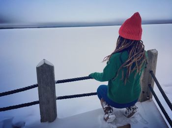 Rear view of mature woman looking at frozen sea while crouching on pier against sky