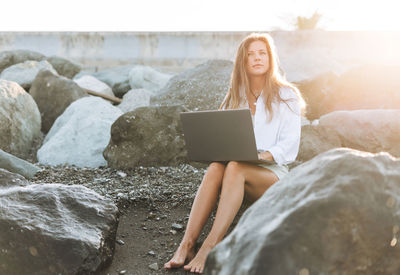 Young carefree freelance business woman with long hair in white shirt working at laptop on seashore 