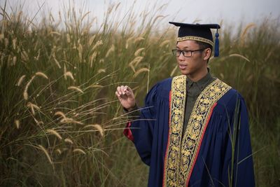 Young man in graduation gown standing on field against sky