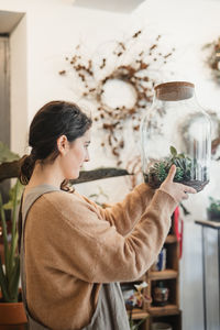 Side view of female in casual clothes holding big transparent glass terrarium closed with cork lid and looking at small green succulents inside while standing against blurred interior of modern flower shop