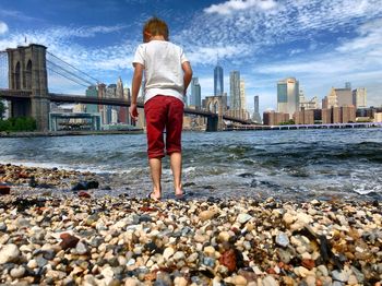 Rear view of boy standing against brooklyn bridge over east river in city