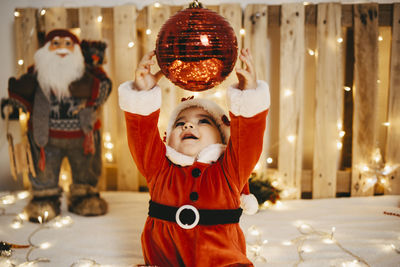 A baby girl disguised as santa claus trying to catch a christmas ball