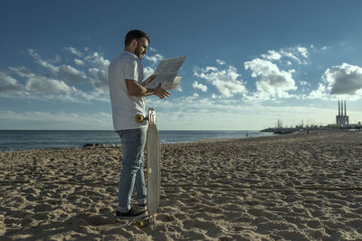 Side view of mid adult man reading map while standing at beach against blue sky during sunny day