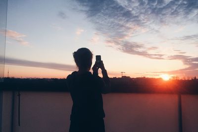 Rear view of woman photographing by river against sky during sunset