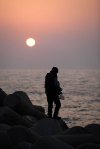 Silhouette man standing on groyne by sea against sky during sunset