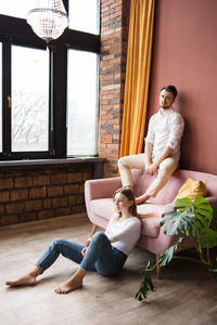 Man sits on sofa's back and look at camera, woman sits on floor near.