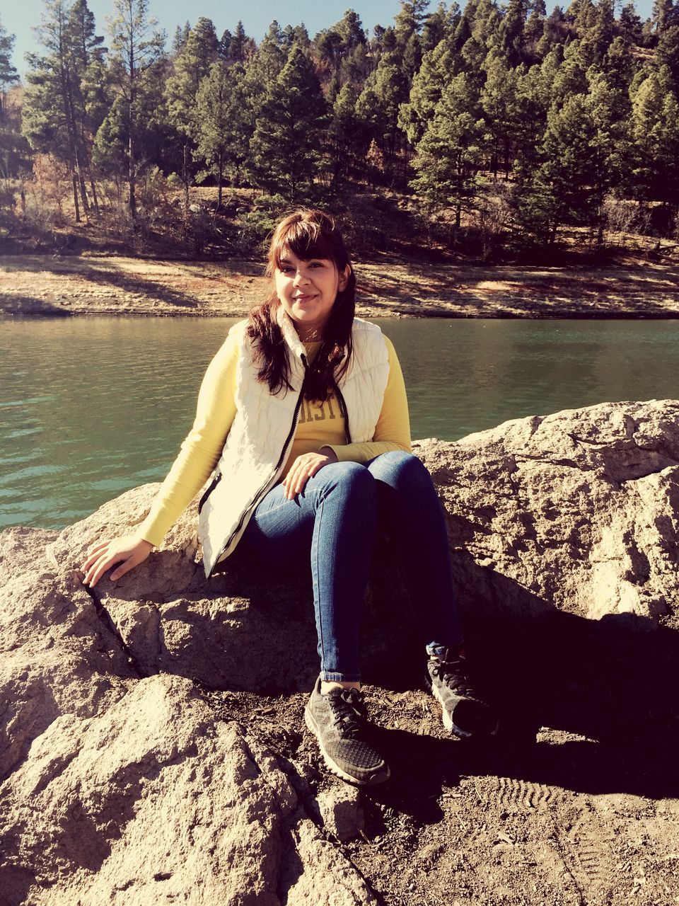 sitting, real people, young women, water, young adult, full length, one person, lake, casual clothing, nature, leisure activity, rock - object, day, outdoors, tree, lifestyles, smiling, looking at camera, beauty in nature, portrait, animal themes, mammal, people