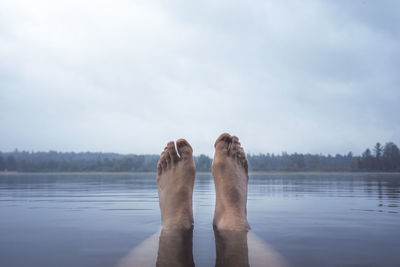 Low section of person relaxing in lake against cloudy sky