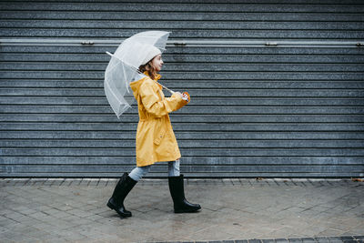 Girl wearing raincoat and jump boot holding umbrella while walking on sidewalk