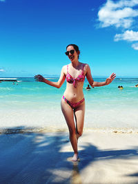 Portrait of young woman with arms raised standing at beach against sky