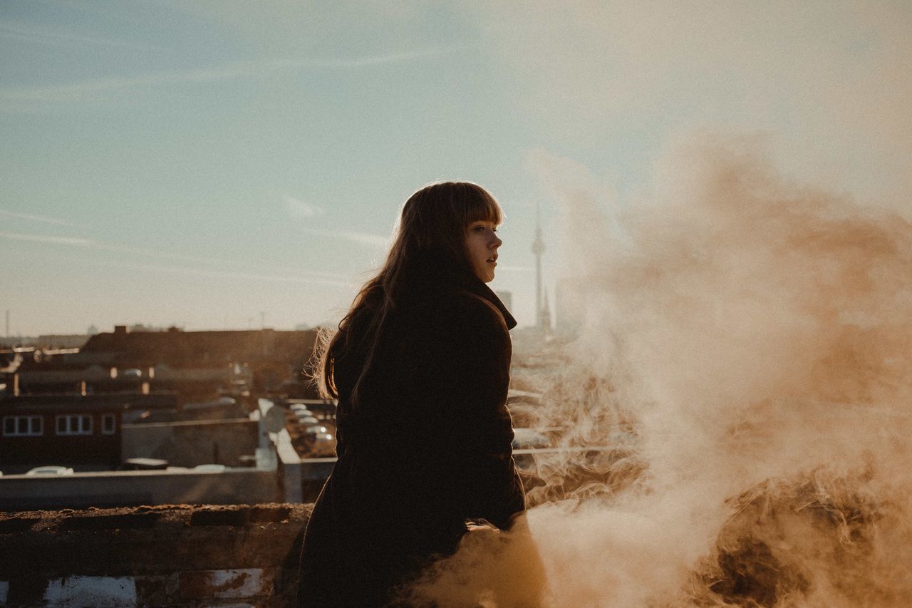 one person, smoke - physical structure, side view, standing, real people, young adult, architecture, sky, three quarter length, lifestyles, young women, casual clothing, built structure, nature, leisure activity, smoke, looking, outdoors, environment, hairstyle, contemplation