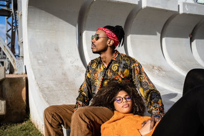 Black man sitting while woman with curly hair lies down in his lap looking away both wearing stylish outfits on concrete bench