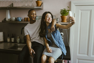 Happy young woman taking selfie with friend sitting on kitchen counter at home