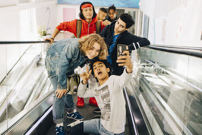 Happy male friends taking selfie with smart phone on moving walkway in shopping mall