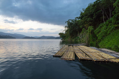 Wooden rafts moored on lake by trees against cloudy sky