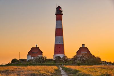 Lighthouse on field by building against sky during sunset