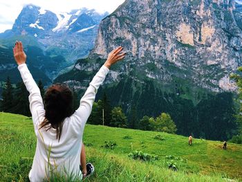 Rear view of woman with arms raised sitting on grassy field against mountains