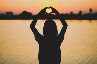 Silhouette woman making heart shape with hands by lake against sky during sunset